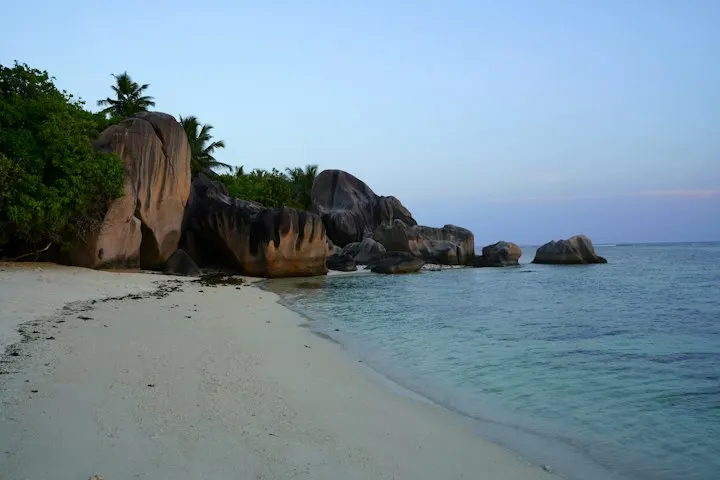 A sandy beach next to a body of water This serene beach scene from the Seychelles showcases the iconic granite boulders that line its pristine shores, gently ki